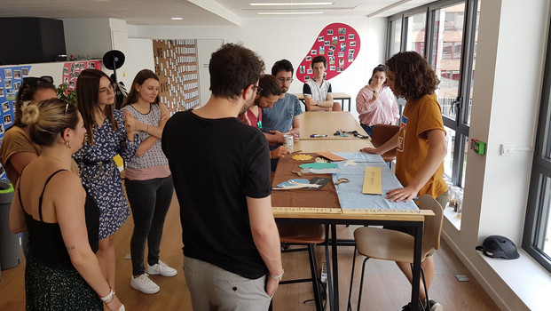 Animation sensibilisation au compostage et au tri des déches organiques par OuiCompost Une séance de sensibilisation de compostage animée par OuiCompost dans les bureaux d'un client. Un groupe de collaborateurs se tiennent autour d'une table pour écouter les explications à propos du tri des déchets alimentaires et le compostage urbain. Sur la table, on voit les photos et les jeux ludiques proposés aux participants de l'atelier.