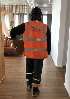 Collecte de biodéchets dans les bureaux par OuiCompost Un collecteur de OuiCompost rentre dans les bureaux d'un client pour collecte les déchets alimentaires de bureau. Le collecteur de OuiCompos porte un gilet orange avec un grand logo de OuiCompost sur le dos et transporte un seau à biodéchets pour remplacer un bac plein qui s'apprête à collecter..