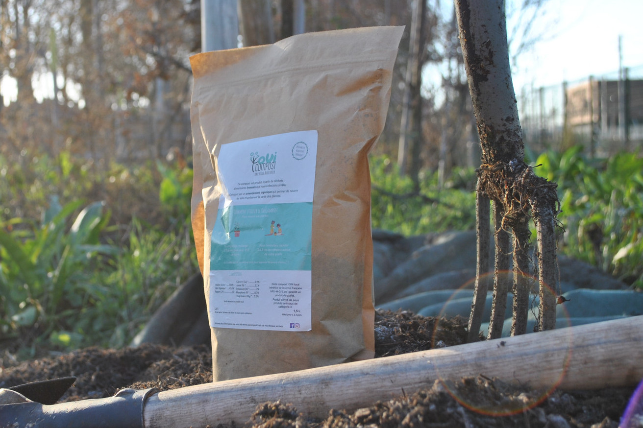 Pochon de compost OuiCompost Un pochon de compost de 1kg avec une étiquette de OuiCompost es posé sur le sol à côté d'une fourche plantée dans le sol. C'est le pochon destiné à la vente de compost produit par OuiCompost à partir des déchets organiques collectes à Lyon.